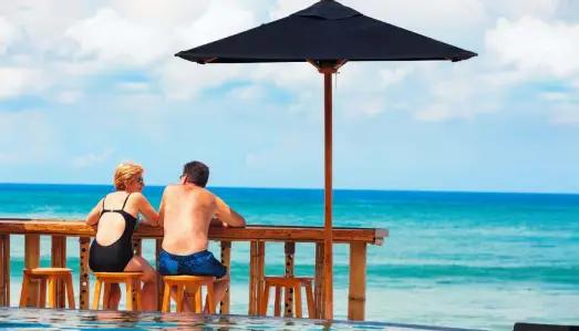 Retired couple strolling along a sunlit Mediterranean promenade, surrounded by palm trees and tranquil sea views.